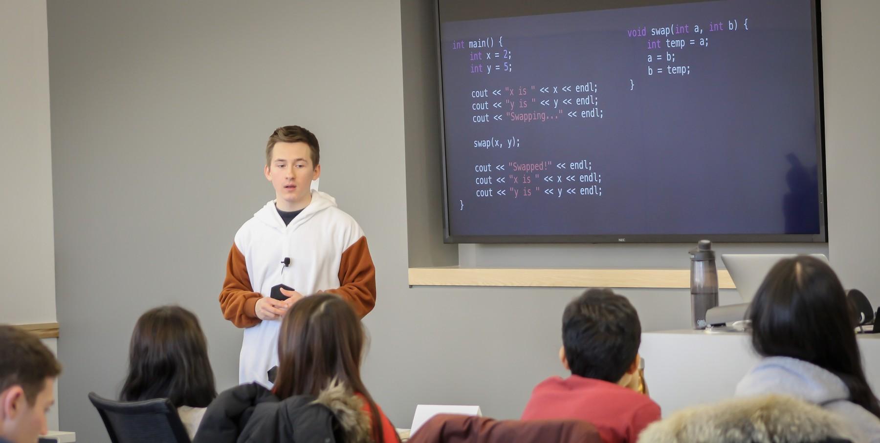 Maxim teaching a programming lecture to a class, C++ code projected on the screen behind him